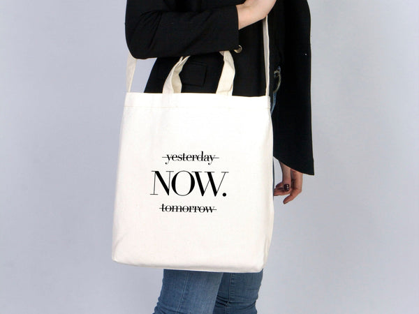 Person holding a tote bag with 'yesterday NOW tomorrow' text on a plain background