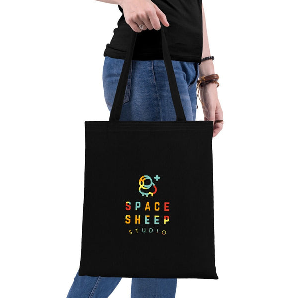 Black tote bag with 'Space Sheep Studio' logo held by a person on a white background