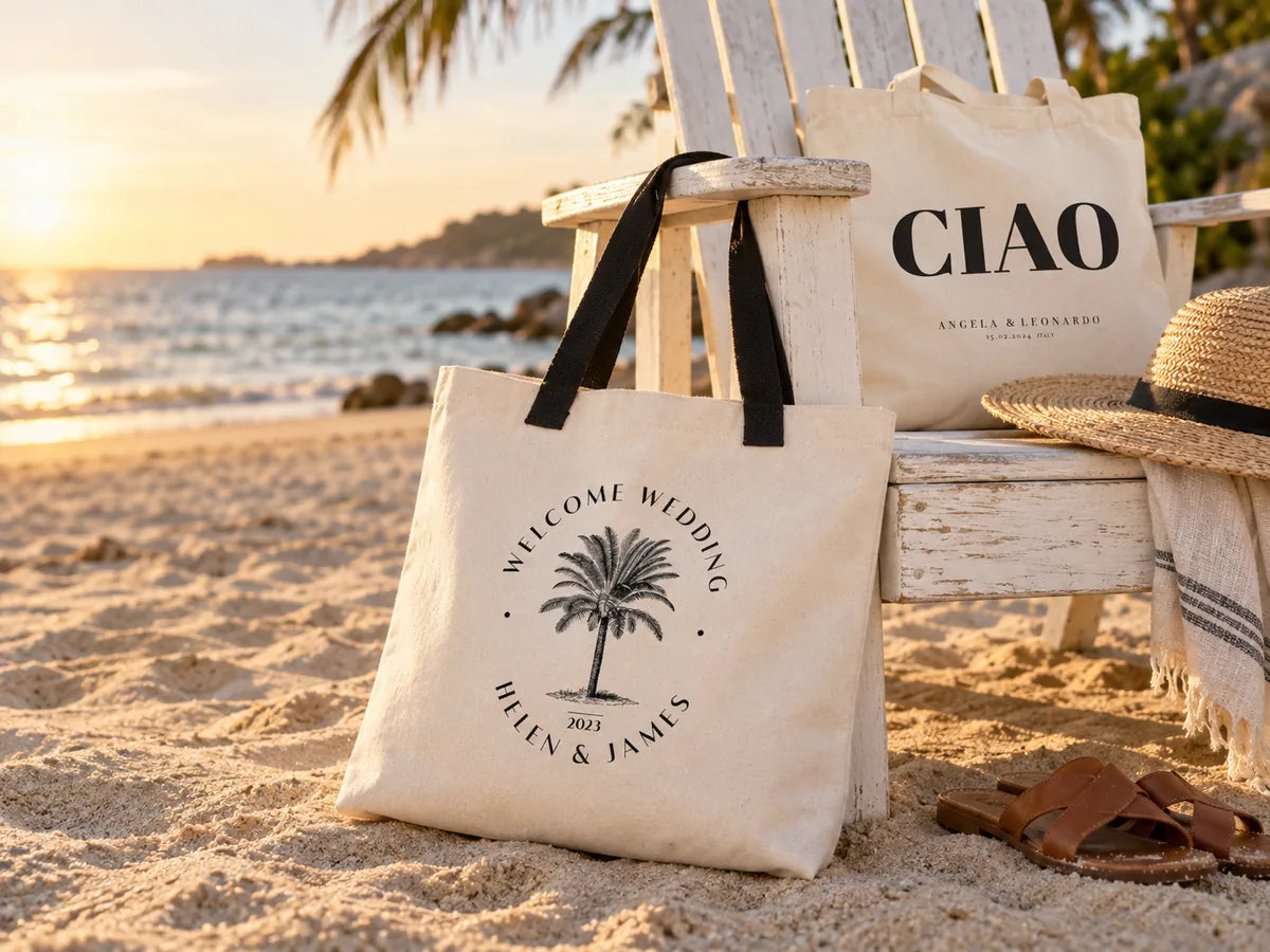 Grote canvas tas ‘Welcome Wedding Helen & James’ met palmboomembleem en zwarte hengsels, leunend tegen een witte Adirondack-stoel op het strand in het gouden uur, met daarachter een tas ‘CIAO’, een strohoed en leren sandalen
